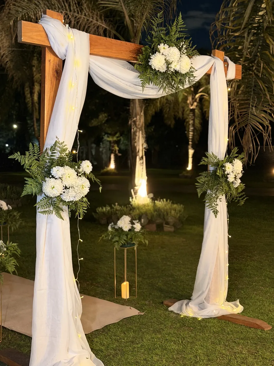 Arco de ceremonia de madera con gasa blanca y flores, iluminado de noche