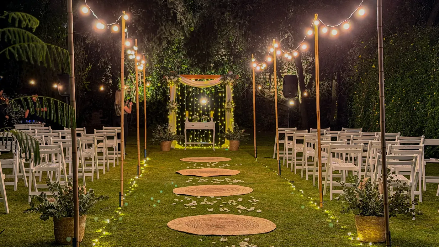 Jardín de bodas de noche con guirnaldas de luces sobre el pasillo de ceremonia y sillas blancas