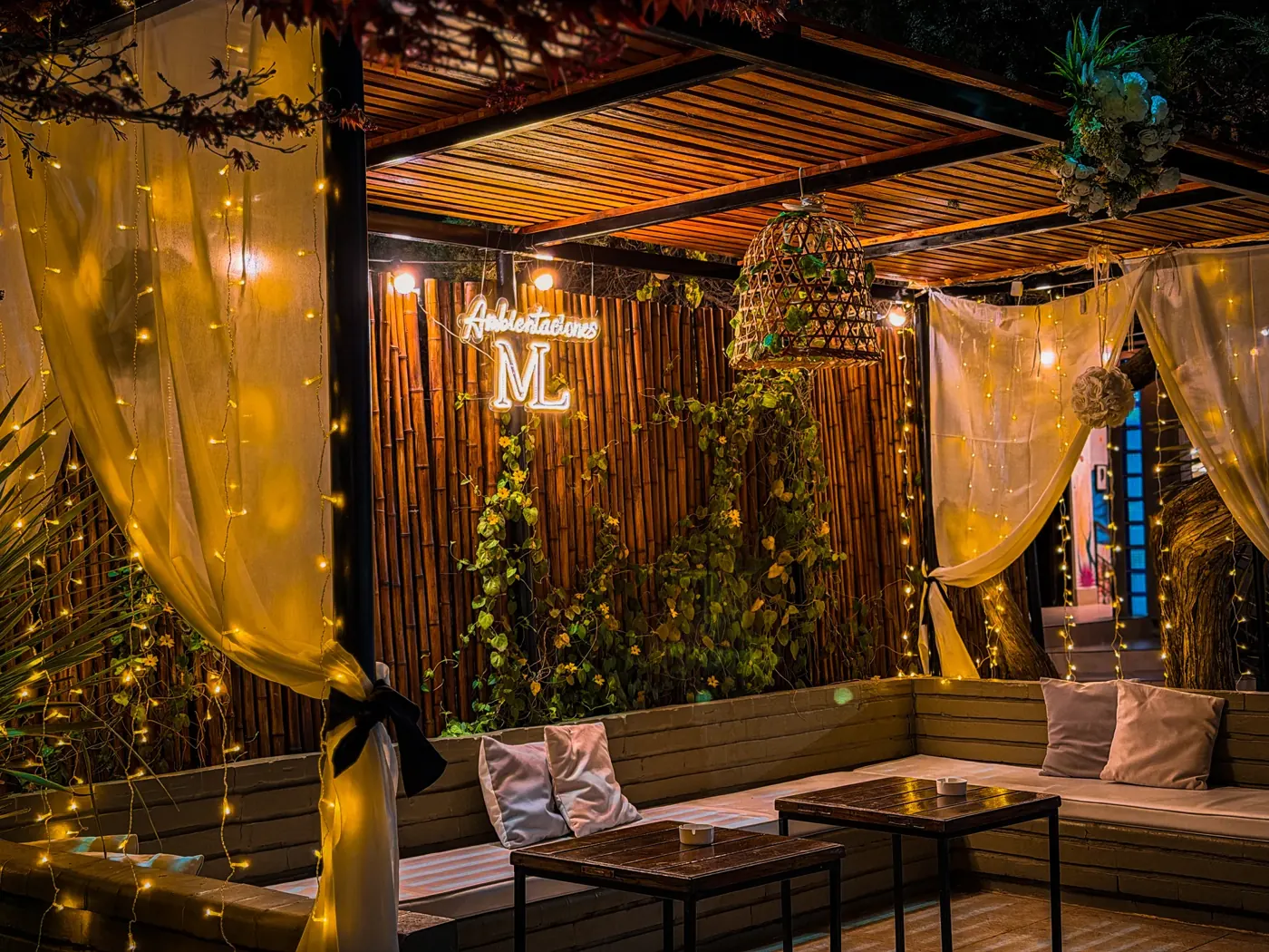 Rincón living bajo pérgola con cortinados, luces y cartel con inicial